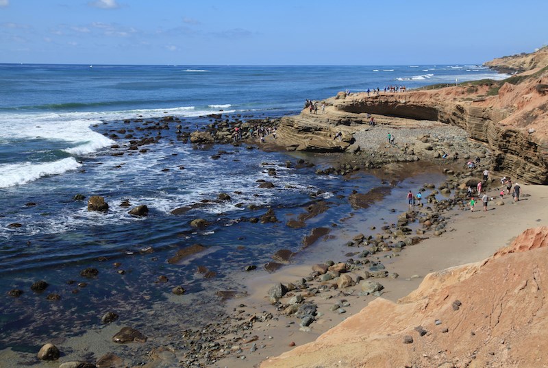 Several people walk on a sandy beach and sandstone cliff near the edge of a large body of water. White waves roll into scattered textured rocks along the beach.